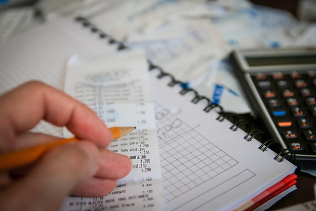 A hand with a pencil reviewing receipts over a notebook and calculator